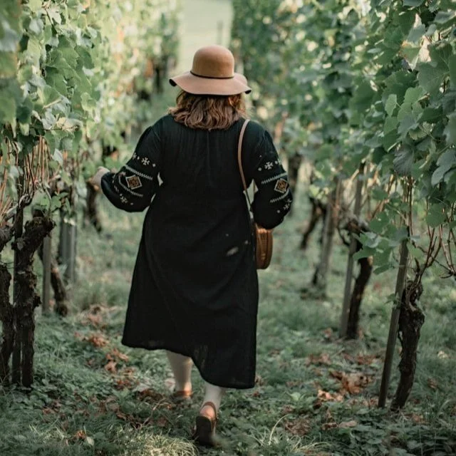 A woman wearing a sunhat, crossbody bag, and sandals walks through a vine row.