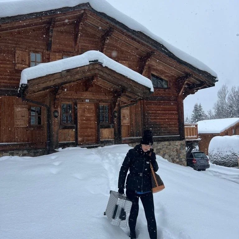 A woman carries a suitcase out of a snow covered ski chalet.
